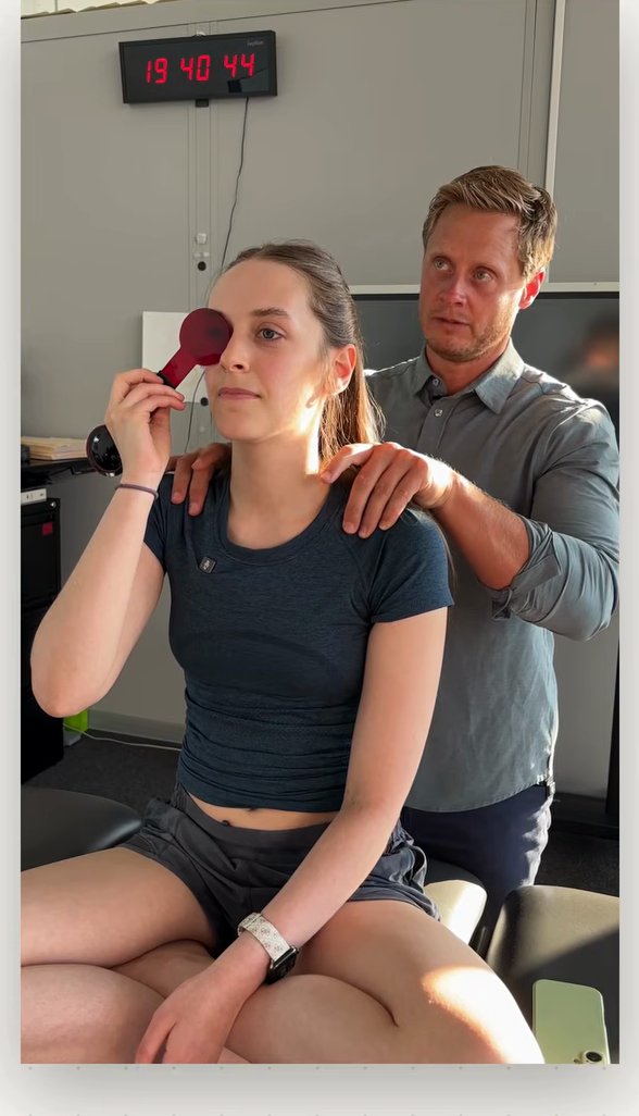 Patient holding a red lens over one eye while the clinician stands behind her providing light feedback through the neck